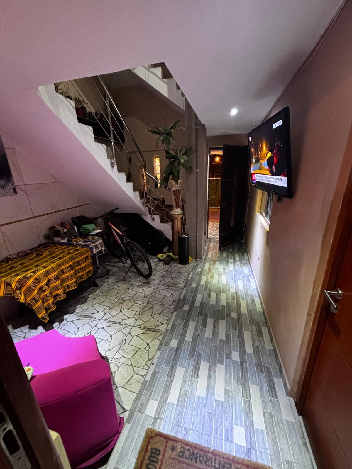 Un pasillo estrecho con una escalera que sube a un piso superior, un televisor montado en la pared y un suelo con diseño.