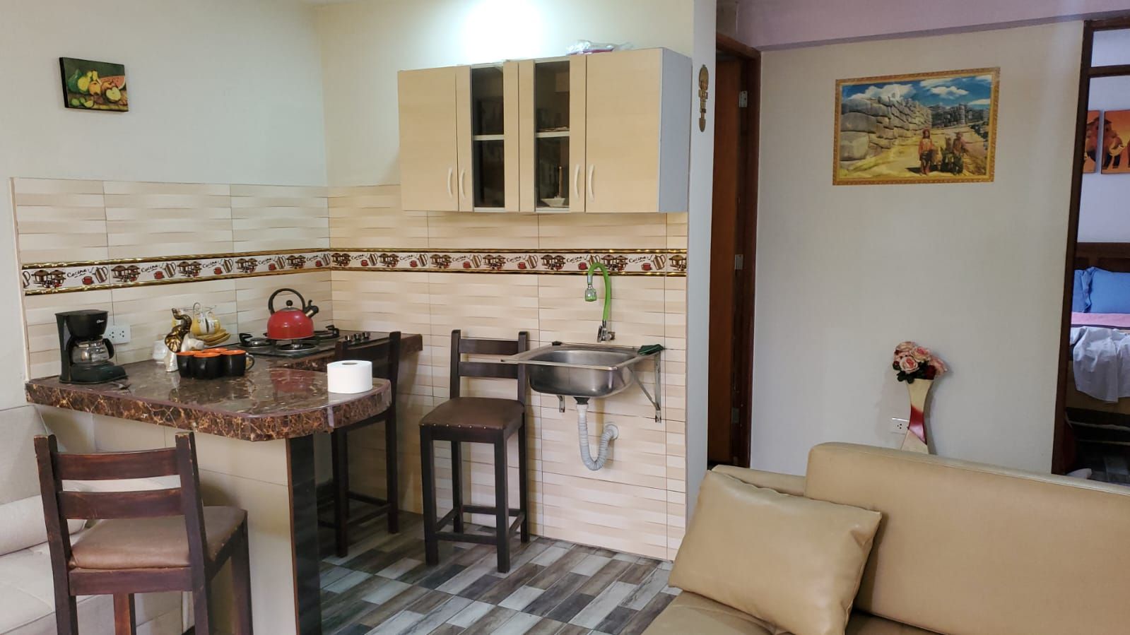Cocina compacta con encimera, lavamanos y armarios. Cuenta con un comedor con sillas y un sofá.