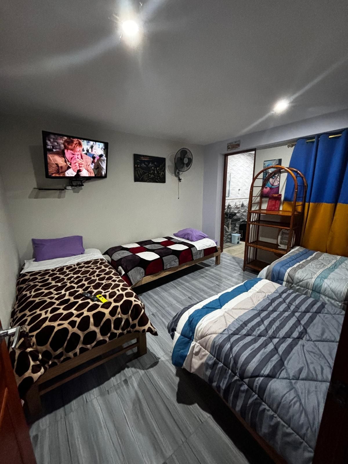 Una habitación con dos camas, un televisor en la pared, un ventilador y una ventana con cortinas.