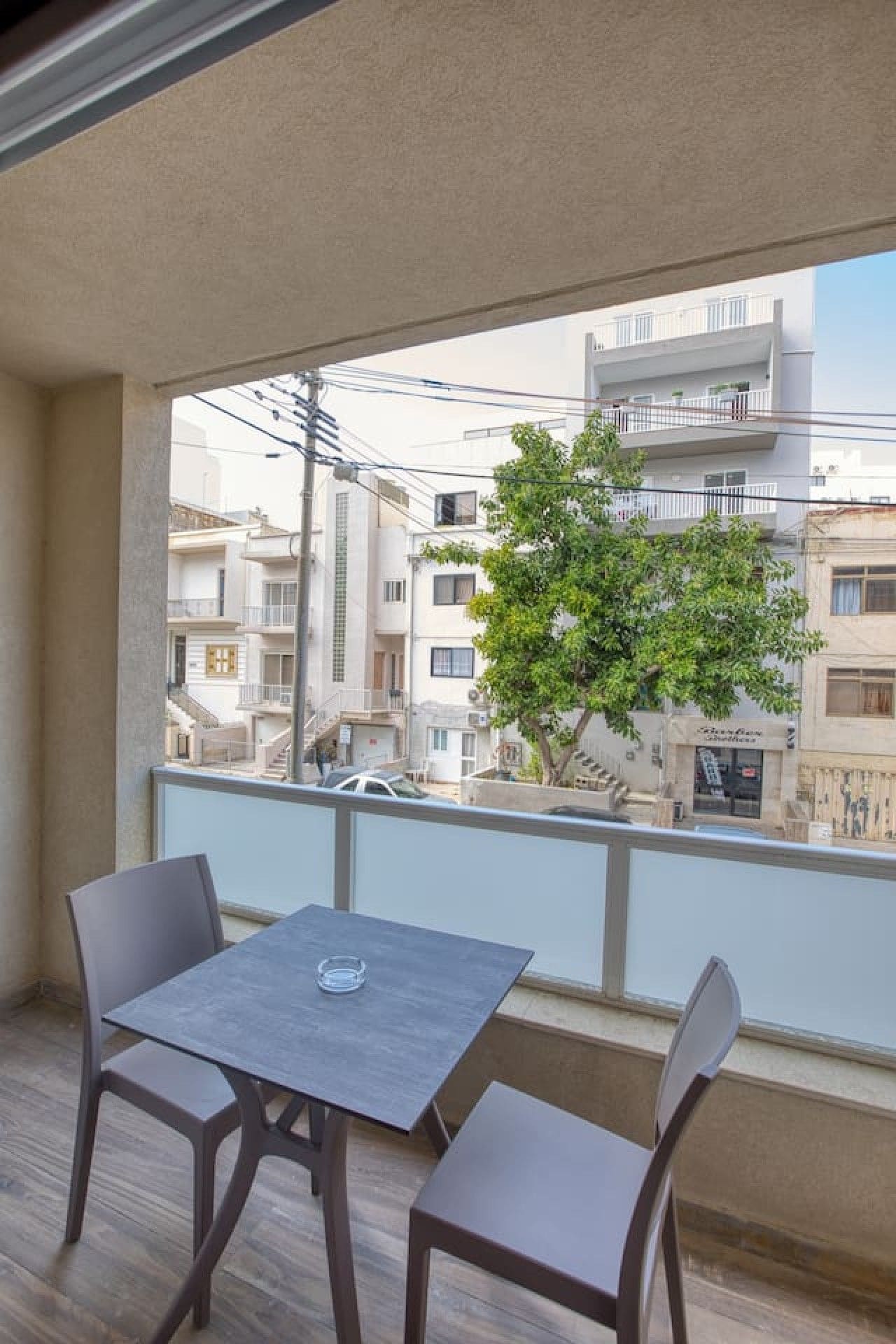 A balcony with a table and chairs, overlooking a residential area with buildings and a tree.
