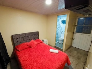 A bedroom with a bed, a nightstand, a bathroom with a toilet, and a door. - La Cachaca (BOGOTÁ)