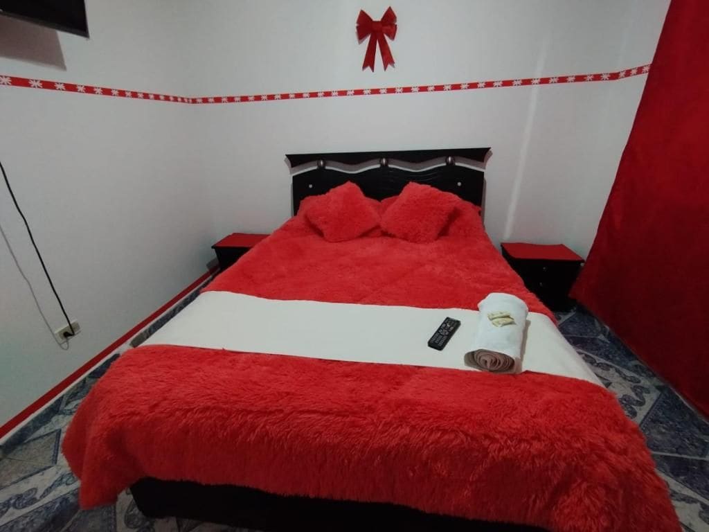 A bed with a red and white bedspread, a remote control, and a rolled towel on the side.