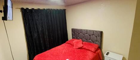 A bedroom with a bed, a headboard, a nightstand, a remote control, and a white trash bin.
