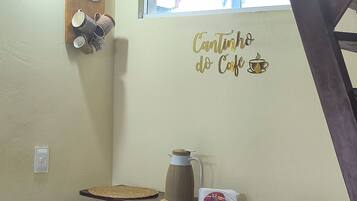 Ett köksutrymme med en hylla där det står muggar och en kaffekanna, samt en väggdekoration. Det finns två pallar och ett fönster ovanför hyllan.