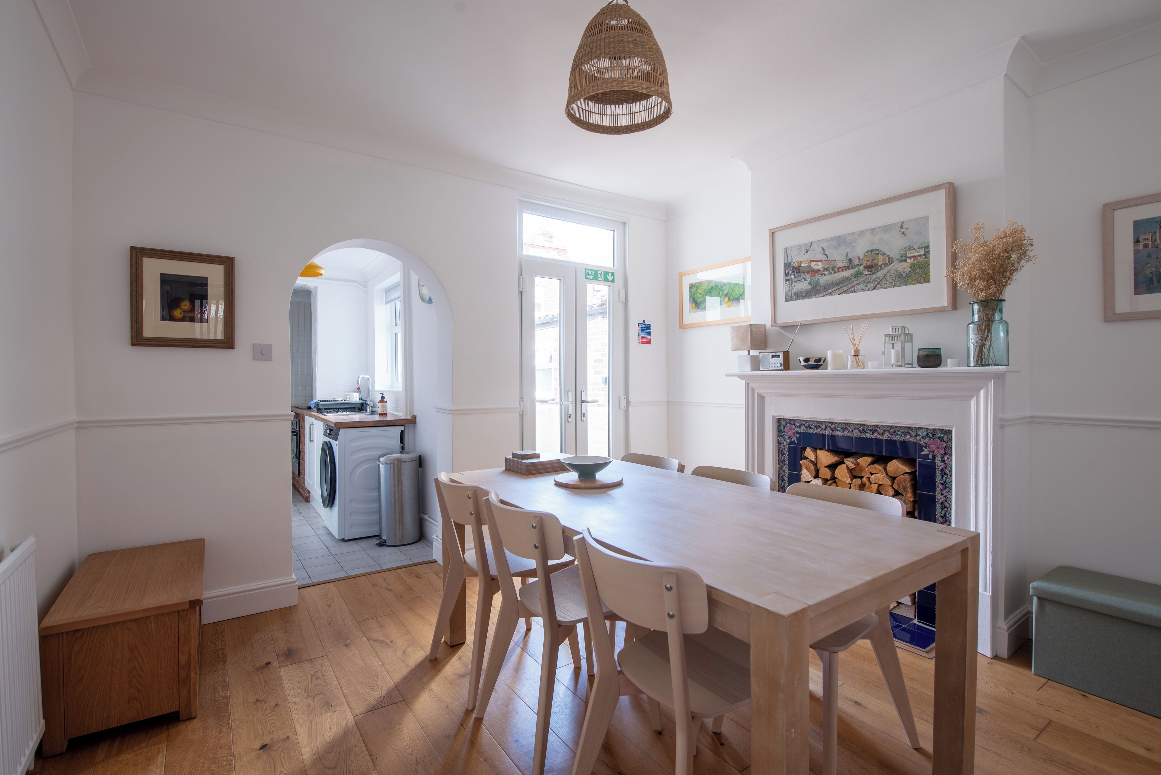 Een eetgedeelte met een houten tafel en stoelen, een keuken op de achtergrond en een open haard met een betegelde omlijsting.