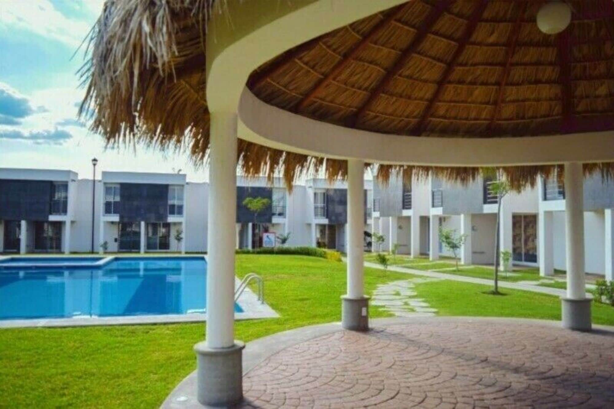 A poolside area with a thatched roof pavilion, a swimming pool, and modern buildings in the background.