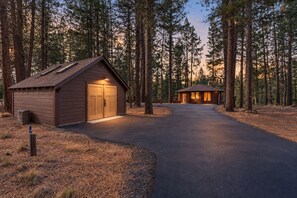 A cabin with a paved driveway leading to a wooden door, surrounded by tall pine trees. - Riverfront Ski Cabin W/ Hot Tub & Dock! (Bend)
