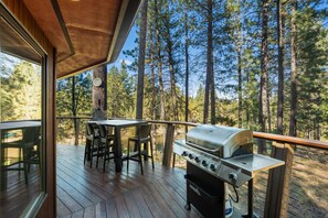 An outdoor deck with a grill, chairs, and a table, surrounded by tall trees. - Riverfront Ski Cabin W/ Hot Tub & Dock! (Bend)