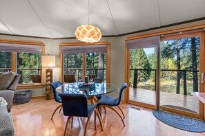 A dining area with a glass table and chairs, a pendant light, and large windows with a view of trees. - Riverfront Ski Cabin W/ Hot Tub & Dock! (Bend)