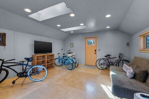 A spacious room with a flat-screen TV, wooden shelving, bicycles, a couch, and a door with a window. - Riverfront Ski Cabin W/ Hot Tub & Dock! (Bend)