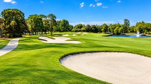 Ein Golfplatz mit Sandbunkern, einem Wasserhindernis und blauem Himmel.