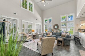 A spacious living room with a high ceiling, large windows, and a modern kitchen area. - Sandestin Cottage on Raven Golf Course: 3 En-Suite BR, Golf Cart, Pool (Miramar Beach)