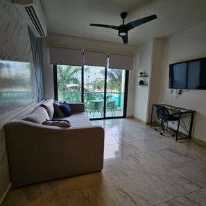 Ein Wohnzimmer mit einer Couch, einem an der Wand montierten Fernseher, einem Deckenventilator und einem Balkon mit Blick auf einen Pool.