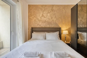 A hotel room with a large bed, a nightstand with a lamp, and a view of the outside through a window. - Sleek 1BR Apartment in Pietà with Balcony (Tal-Pietà)