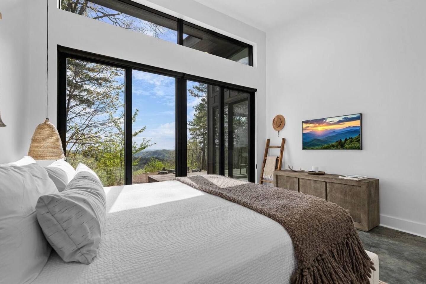 A bedroom with a large bed, a view of the outdoors, a wooden dresser, and a painting on the wall.