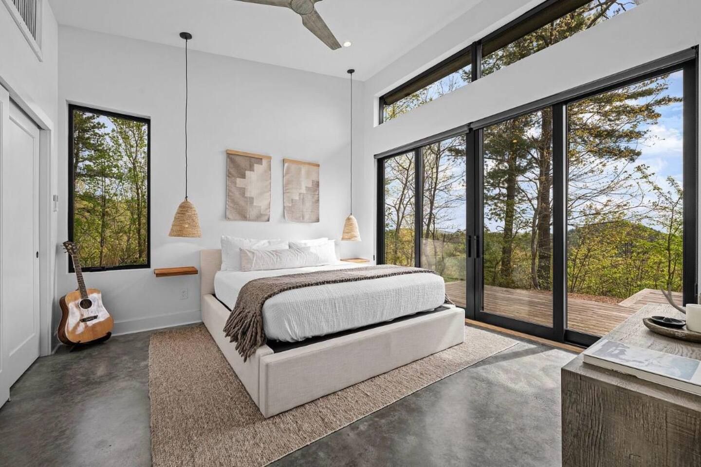 A modern bedroom with a large bed, a guitar, and a view of trees through a sliding glass door.