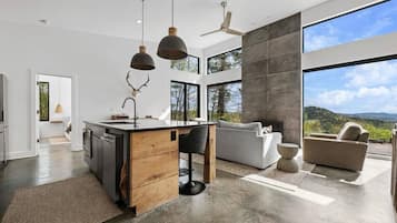 Una cocina moderna con una isla central, una zona de comedor con un sofá y ventanas amplias que ofrecen vistas al exterior.