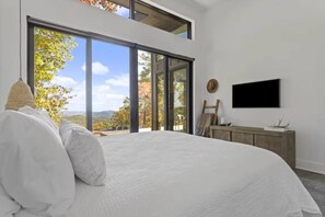 A bedroom with a large bed, a television mounted on the wall, and a view of the outdoors through large windows. - Luxury Glass Cabin + Hot Tub near Blowing Rock (Lenoir)