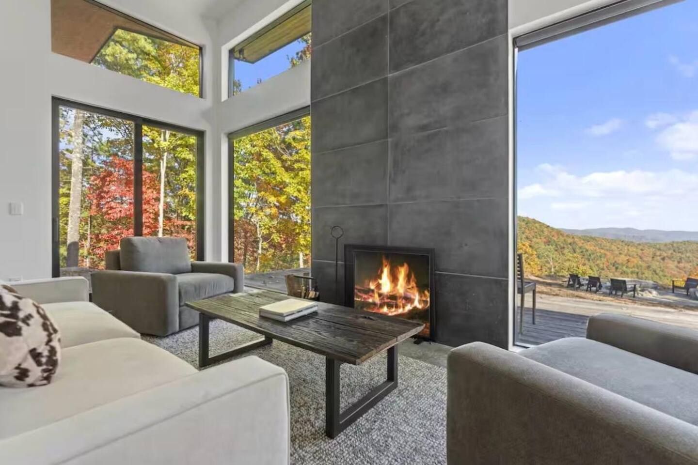 A modern living room with a fireplace, large windows, and a view of autumn foliage.