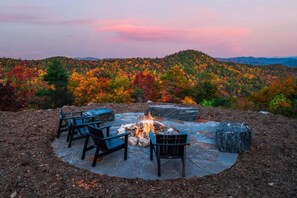 An outdoor seating area with a fire pit surrounded by chairs, set against a backdrop of autumn foliage and distant hills. - Luxury Glass Cabin + Hot Tub near Blowing Rock (Lenoir)