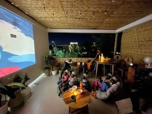 Ein Innenraum mit einer Projektionsleinwand, die eine Landschaft zeigt, einer Gruppe von Personen, die auf dem Boden sitzen, und einem Tisch mit Gegenständen darauf.