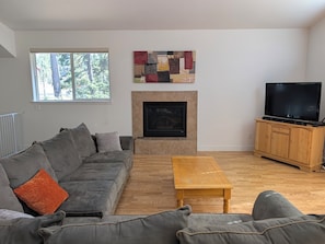 Ein Wohnzimmer mit einer Couch, einem Couchtisch, einem Kamin, einem Fernseher und einem Fenster mit Blick auf Bäume.