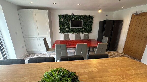 Un comedor con una mesa de madera, seis sillas, un mantel rojo y un televisor fijado a la pared.