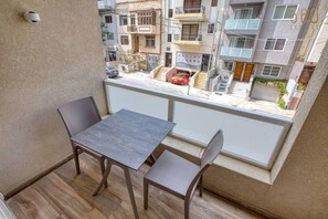 Un balcone con tavolo e sedie, affacciato su una strada con edifici e un'auto parcheggiata.