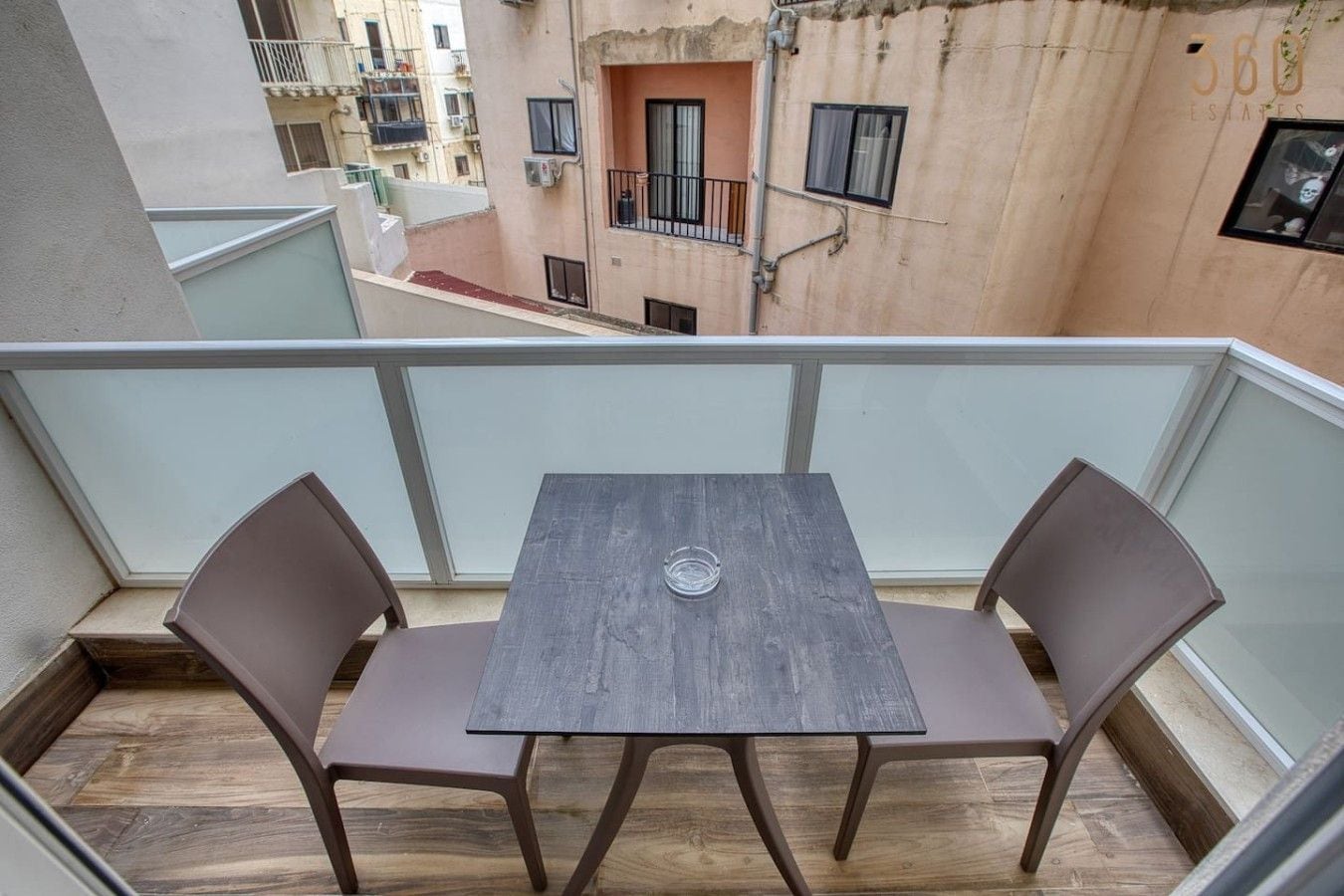 A balcony with a table and two chairs, overlooking a residential area with buildings.