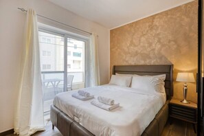 Ein Hotelzimmer mit Bett, Nachttisch, Lampe und Blick nach draußen.