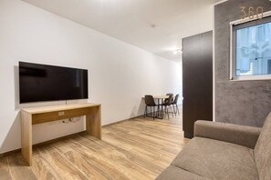 Ein modernes Wohnzimmer mit einem Flachbildfernseher auf einer Holzkonsole, einem grauen Sofa und einem Essbereich mit Stühlen.