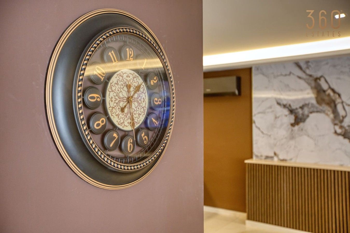 Une horloge murale avec un cadre décoratif, un mur en marbre et un comptoir d’accueil en arrière-plan.