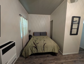 Een slaapkamer met een bed, een nachtkastje, een lamp, een raam met gordijnen, een radiator en een spiegel aan de muur.