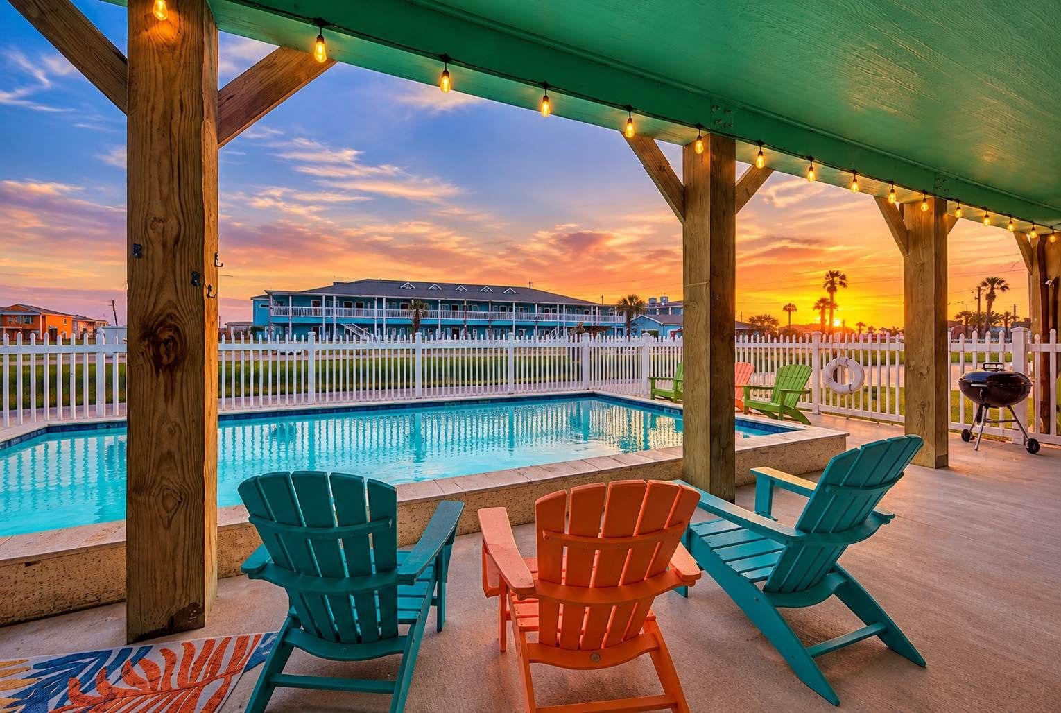 A poolside area with chairs and a barbecue grill, overlooking a pool and a building with a white fence.
