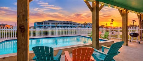 A poolside area with chairs and a barbecue grill, overlooking a pool and a building with a white fence.
