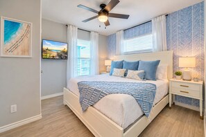A bedroom with a bed, a ceiling fan, a nightstand with a lamp, and a wall-mounted TV. - Coastal Escape | Private Pool + Games | Sleeps 12 (Port Aransas)