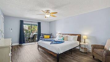 A bedroom with a bed, a ceiling fan, a chair, a bedside table, and a view of the outdoors.