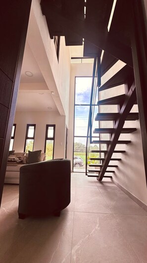 A modern interior with a staircase leading to an upper level, a sofa, and a view of the outdoors. - Sapphire House (La Perla)
