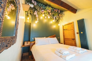 A hotel room with a bed, pillows, a wooden headboard, a wooden door, a radiator, and a wall with hanging lights and flowers.