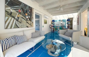 Una sala de estar moderna con suelo de baldosas azules, sofás blancos con cojines a rayas, una mesa de centro de vidrio y un cuadro abstracto grande en la pared.