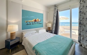 A hotel room with a bed, two bedside lamps, a painting of a person on a hammock, a window with a view of the sea, and a balcony with a chair and umbrella. - Beautiful home in Avola with WiFi (Avola)