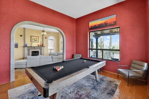 A pool table in a room with red walls, a window with a cityscape view, and a seating area with a sofa and armchairs. - Tucson: Catalina Foothills Retreat (Tucson)