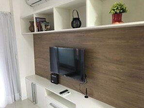 En platt-TV monterad på en vägg med en träpanel, en vit TV-bänk under och en hylla ovanför med dekorativa föremål.