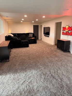 A living room with a sectional sofa, a flat-screen TV mounted on the wall, and a Red Bull sign. - 4-bedroom House in Prime SLC Location Minutes From The U (Salt Lake City)