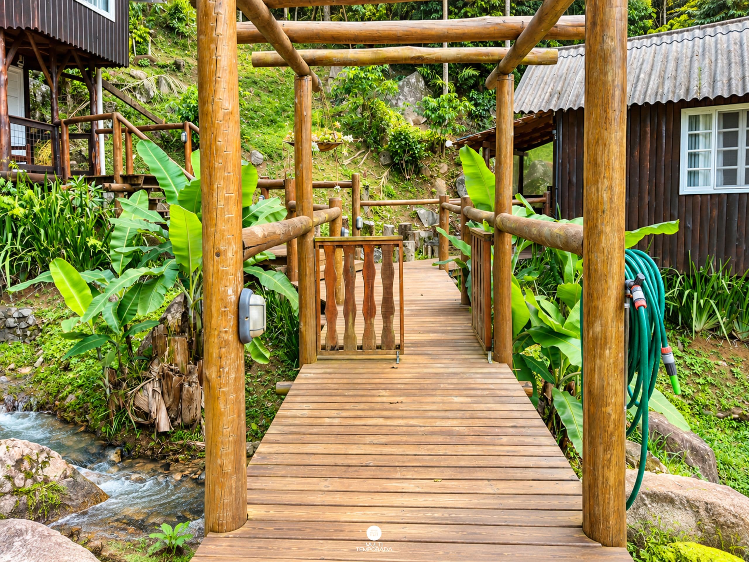 A wooden pathway leading to a wooden structure with a metal gate, surrounded by lush greenery and a small stream.