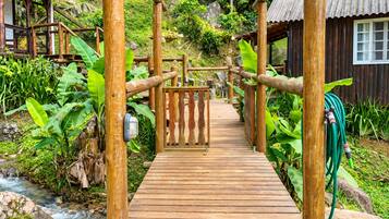 A wooden pathway leading to a wooden structure with a metal gate, surrounded by lush greenery and a small stream.