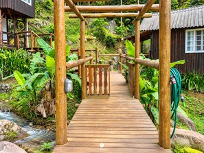 A wooden pathway leading to a wooden structure with a metal gate, surrounded by lush greenery and a small stream.