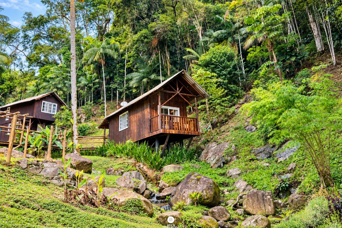 A wooden cabin with a balcony, surrounded by lush greenery and rocks.