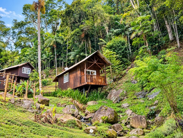 A wooden cabin with a balcony, surrounded by lush greenery and rocks.