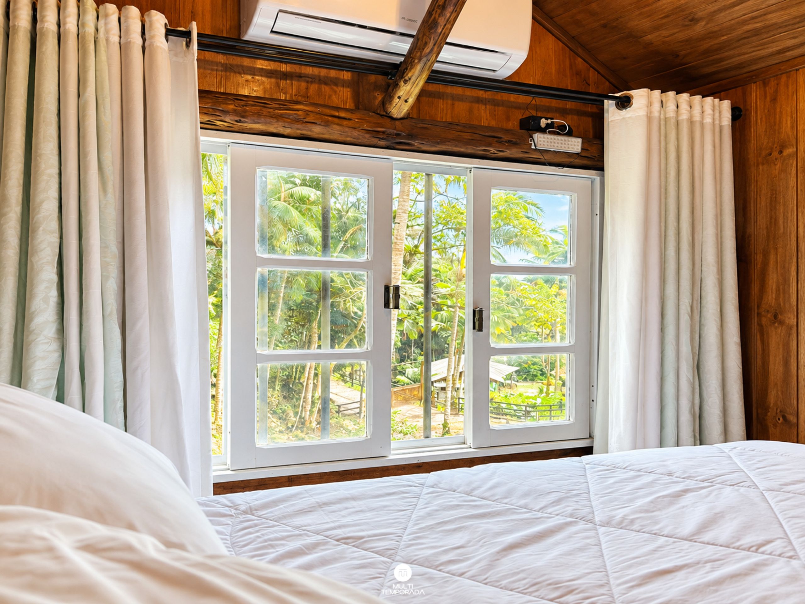 Un dormitorio con un amplio ventanal, techo de madera y una cama con ropa de cama blanca.